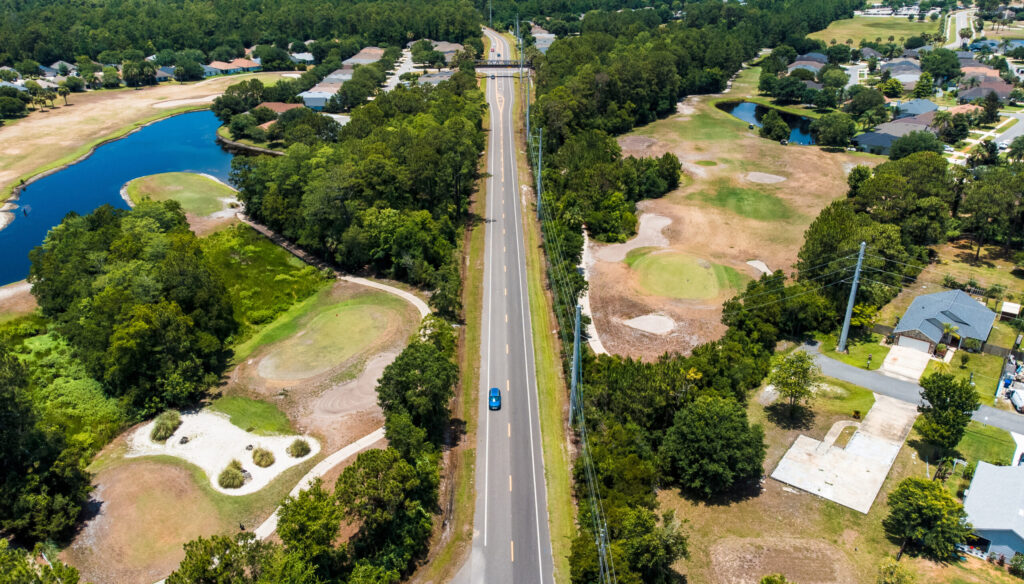 A street runs in between a golf course with trees, a pond and housing development on both sides.