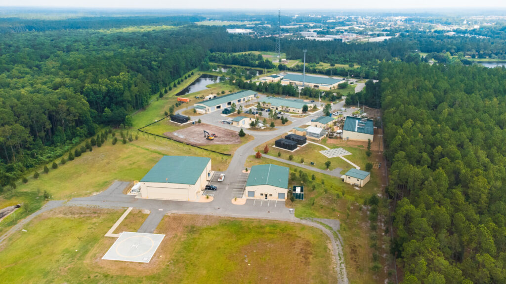 Aerial picture of multiple buildings that are cream colored with green roofs and green space around them. There is dense forest on the right and left and in the background is a metal tower with buildings beyond that.