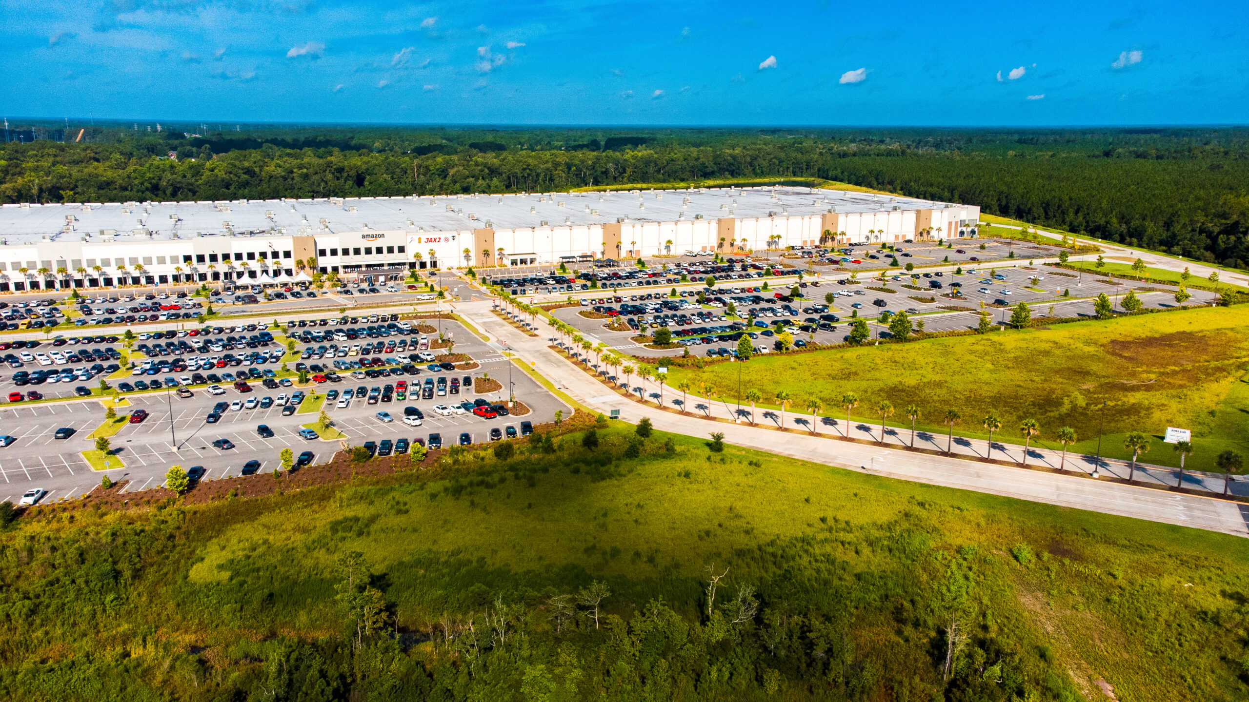 Large white warehouse with parking lots on either side of the road leading up to the building. In the foreground is green space with trees and dense forest surround the building in the background.