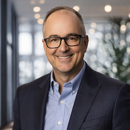 A professional headshot of a smiling man wearing glasses in a blue suit