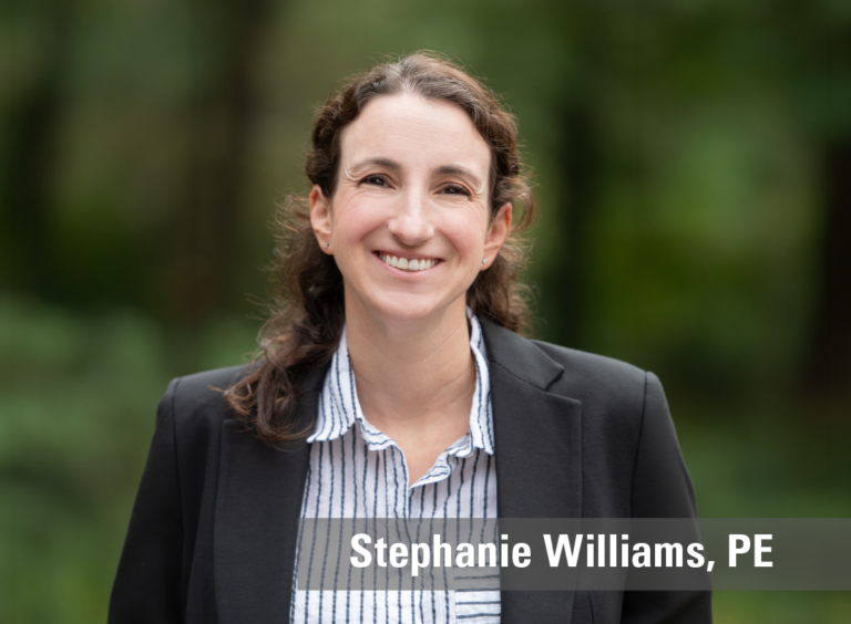 A professional headshot of a smiling woman wearing a grey suit with a striped shirt