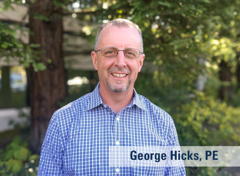 An outdoor photograph of a smiling man wearing a blue plain shirt