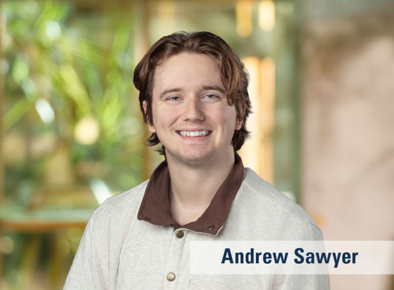 A professional headshot of a smiling young man wearing a tan shirt with a brown collar