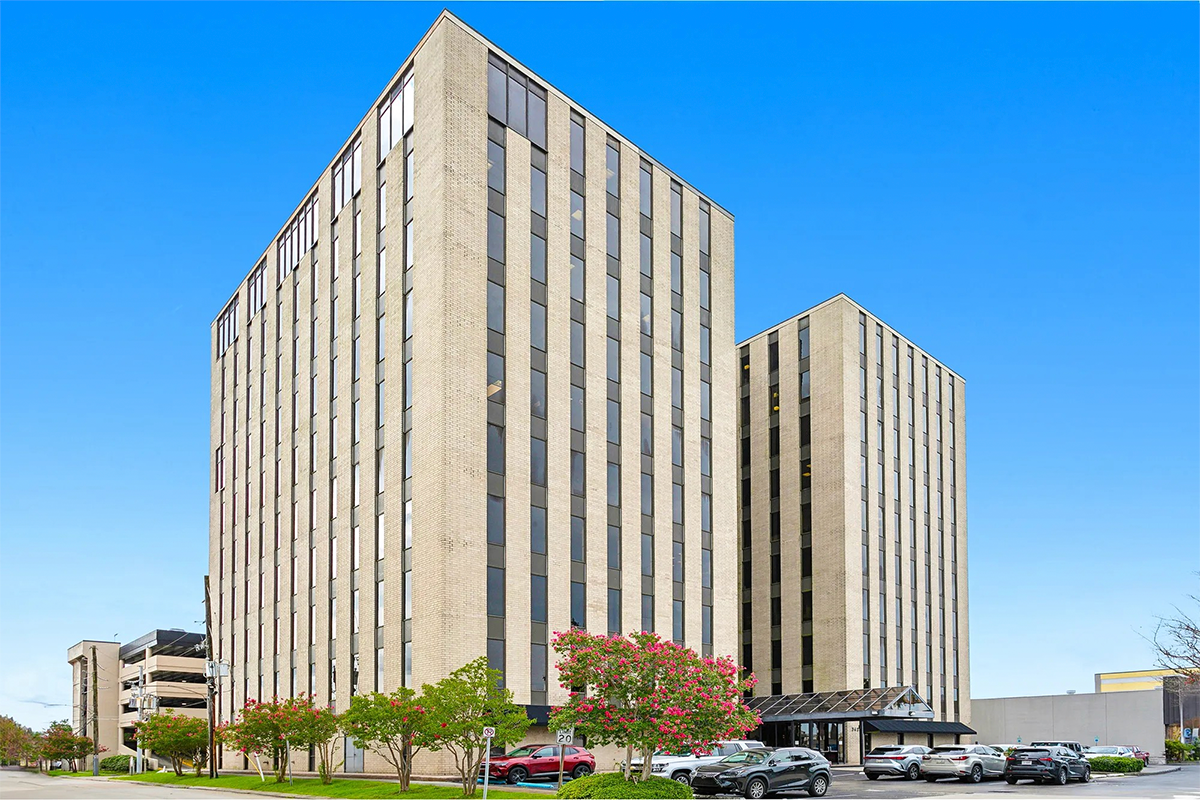 Two large office towers in front of a blue sky with flowering trees and cars
