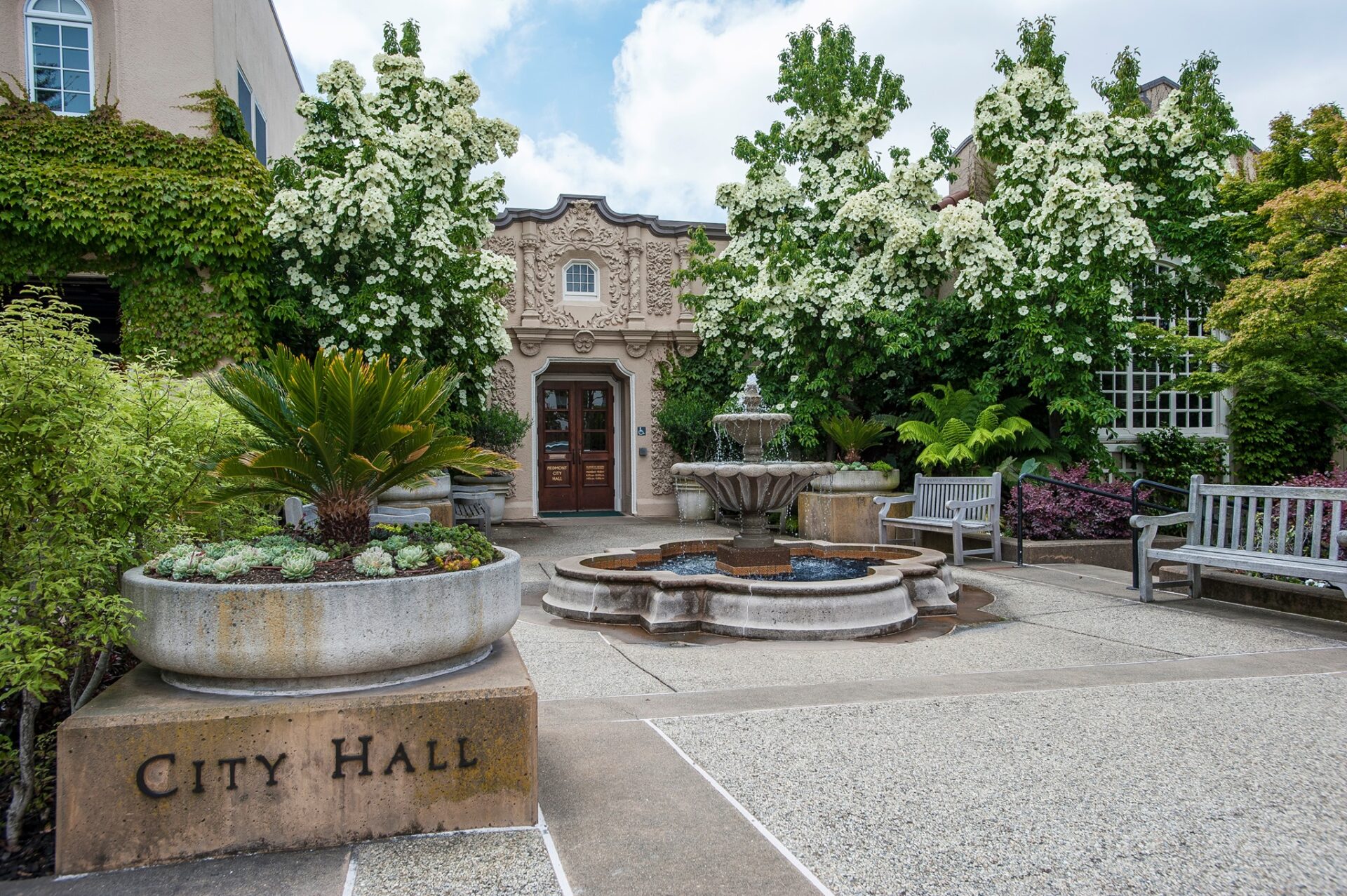 Landscaped city hall courtyard with a fountain, benches, planters, and flowering trees.