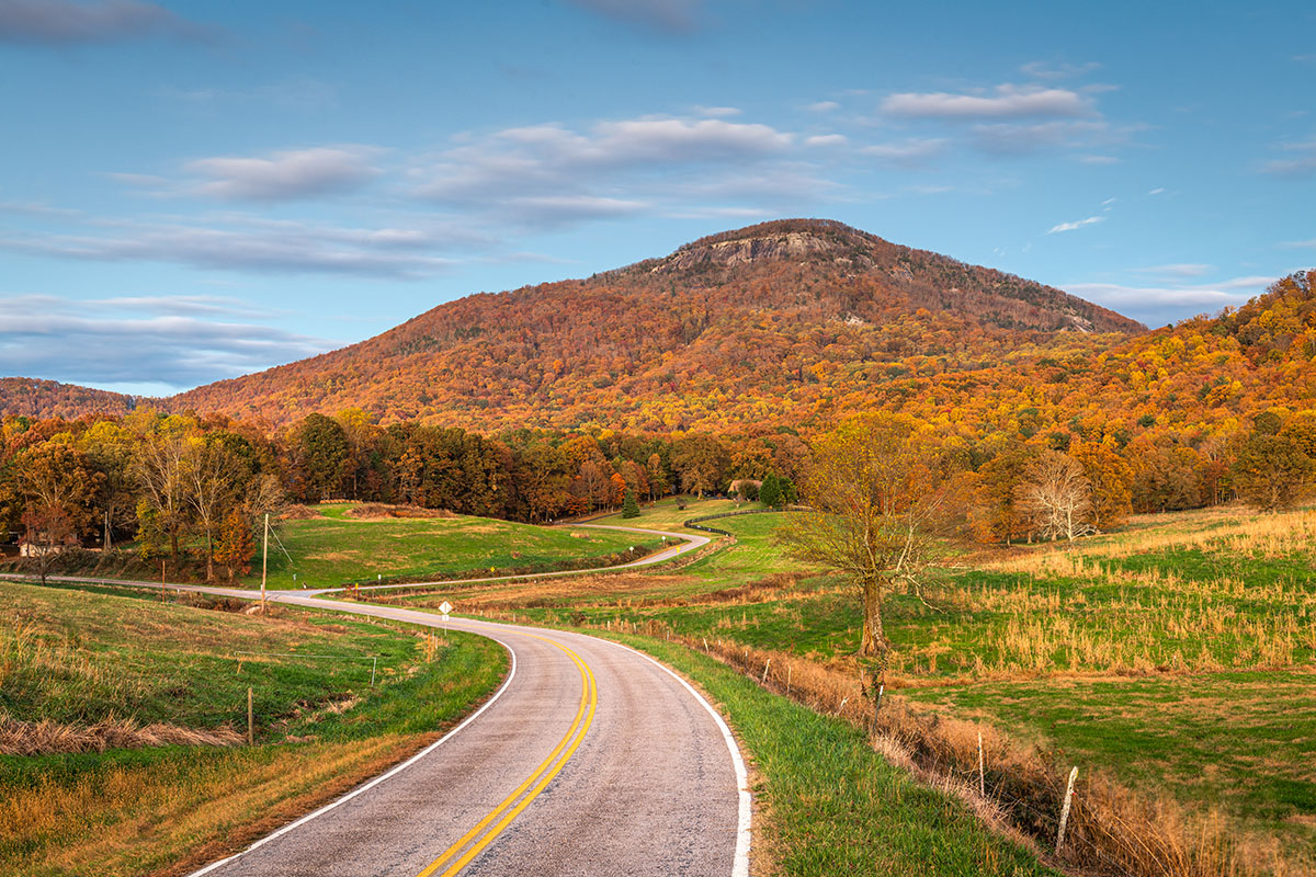 Winding rural road leading toward a forested mountain with colorful fall foliage.